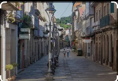 two people walking down a city street with buildings at Un rincón romano in Caldas de Reis