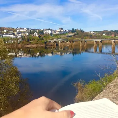 a person reading a book next to a body of water at A Fontana de Luxo Apartamentos in Portomarin